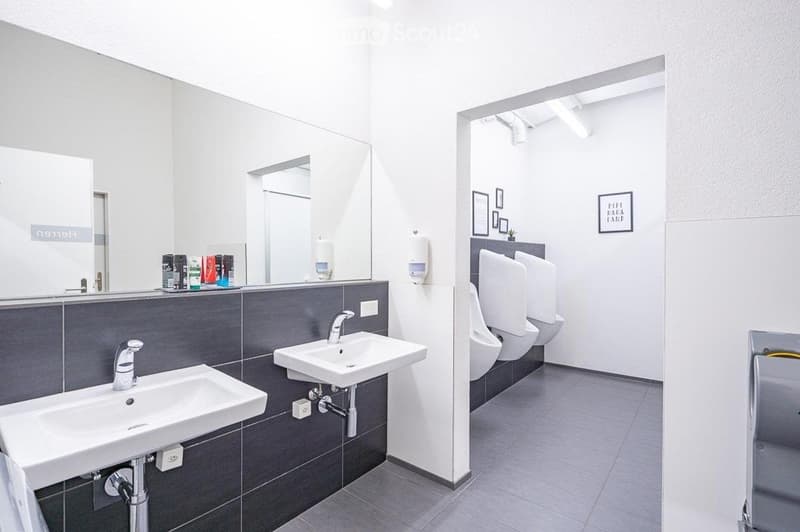 Modern public restroom with two sinks, mirrors, and two urinals. Grey tiled walls and white fixtures.