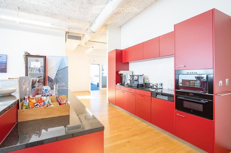 Modern kitchen with red cabinets, stainless steel appliances, granite countertop, and a coffee machine. A refrigerator and a sink are visible, along with a small display of snacks on the counter.