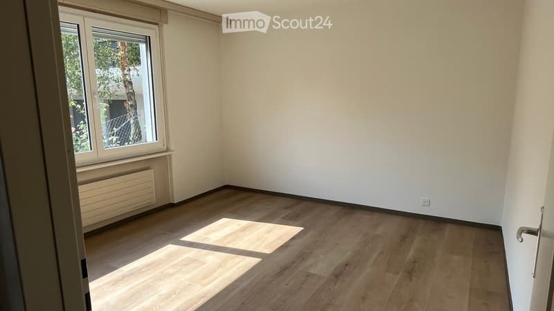 Empty room with light wooden flooring, white walls, and a large window letting in natural light. No furniture or equipment is present.