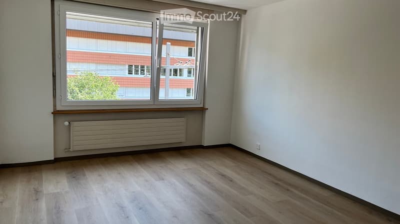 Empty room with wooden flooring, white walls, and a large window with a view of a building and greenery. A radiator is visible below the window.