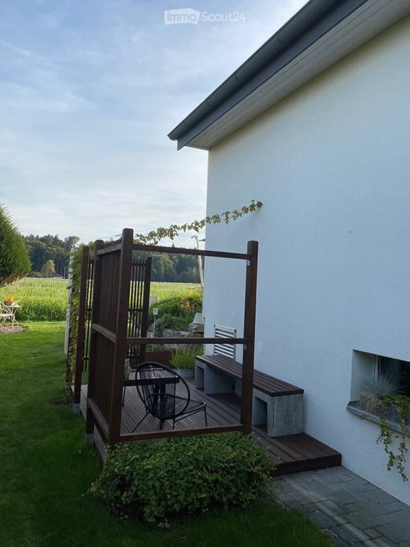 A wooden deck area attached to a house, featuring a bench, a small table, and a chair, surrounded by greenery and a wooden fence.
