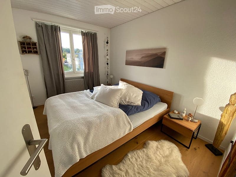 Cozy bedroom with a wooden bed, white bedding, nightstand with lamp, picture frame on wall, and a small shelf. The room has wooden flooring and large windows with curtains.
