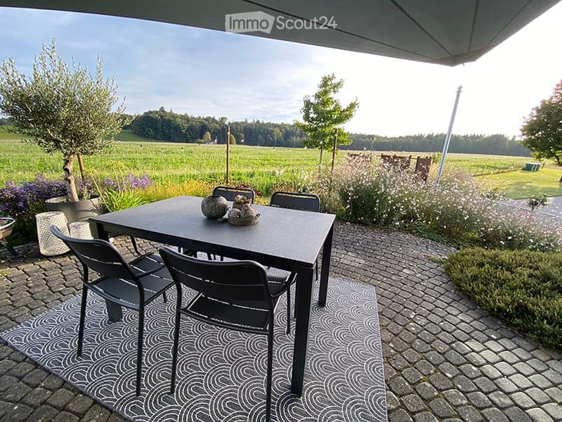 Outdoor dining area with a black table and chairs on a patterned rug, surrounded by potted plants and a scenic countryside view.