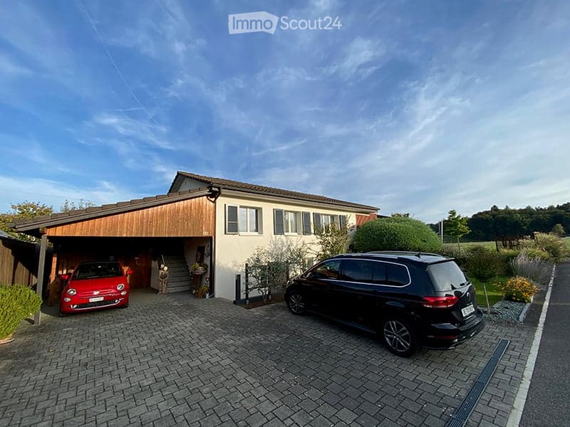 Two-story house with a wooden garage, white walls, multiple windows, and a paved driveway with two parked cars.
