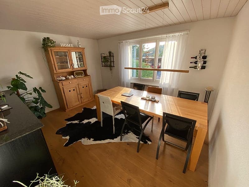 Bright dining area with wooden dining table, chairs, a cabinet with glass doors, and a wine rack on the wall. A cowhide rug is on the floor, and a plant is placed on the cabinet.