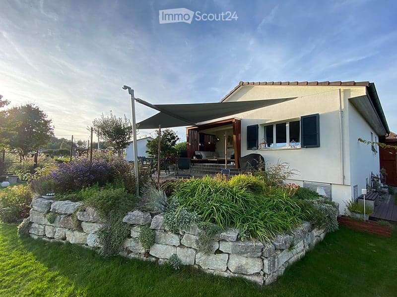 A well-maintained garden area with a stone wall, various plants, and a covered outdoor seating area attached to a house.
