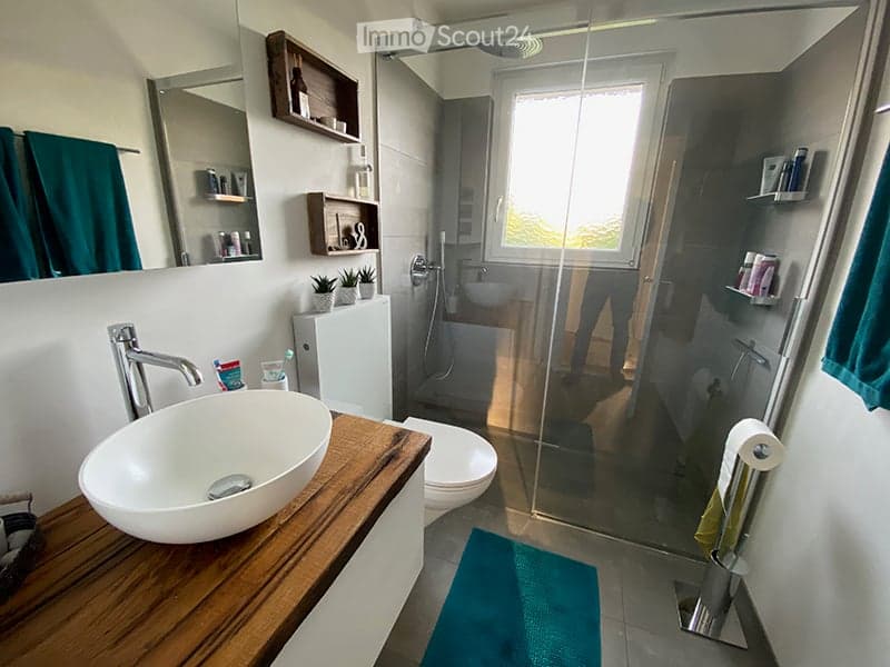 Modern bathroom with a wooden vanity, white sink, chrome faucet, toilet, glass-enclosed shower, and shelves with toiletries and decorative items.