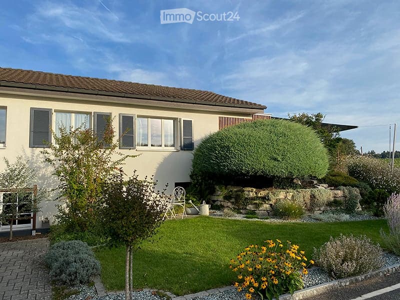 Single-story house with white exterior, multiple windows with shutters, and a well-maintained garden with lawn, shrubs, and flowers.