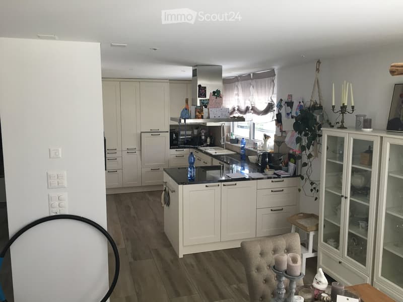 Modern kitchen with white cabinets, black countertops, and wooden flooring. Features a sink, faucet, oven, refrigerator, and a small dining area with a table and chair.