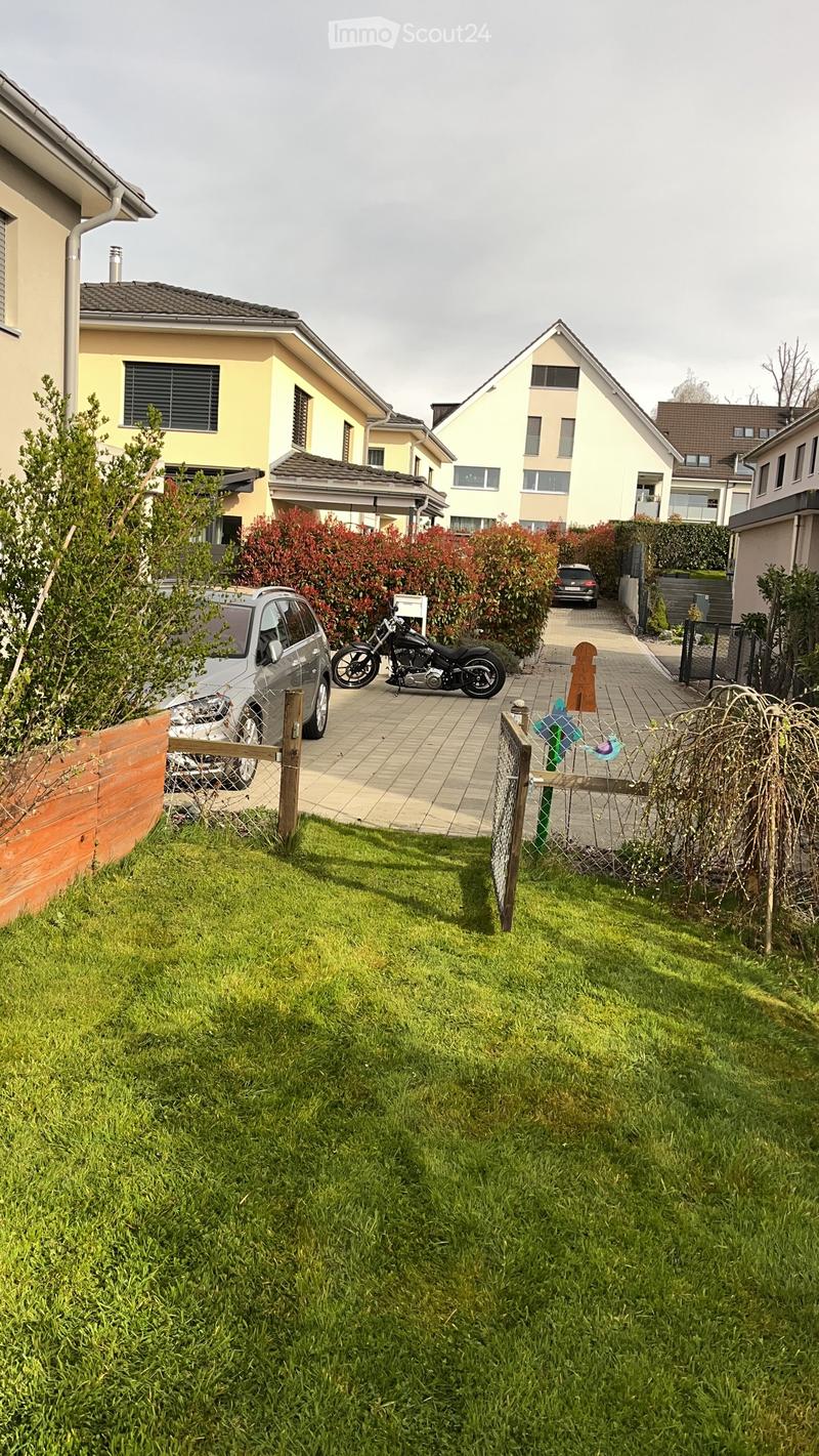A well-maintained garden with a green lawn, a wooden fence, and a small pathway leading to a driveway with parked vehicles, including a motorcycle and a car.