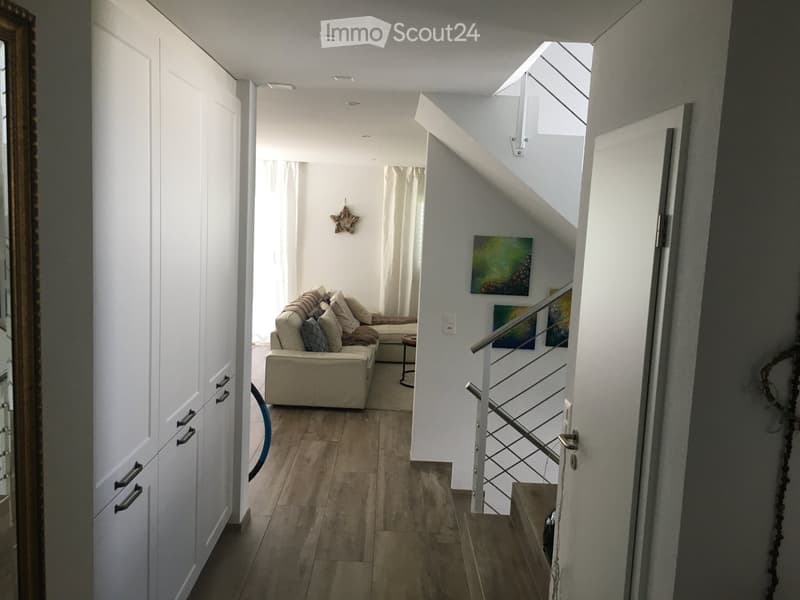 Modern hallway with a small living area featuring a sofa, a small round table, and a mirror on the wall. The hallway has white walls, wooden flooring, and a staircase with a metal railing.