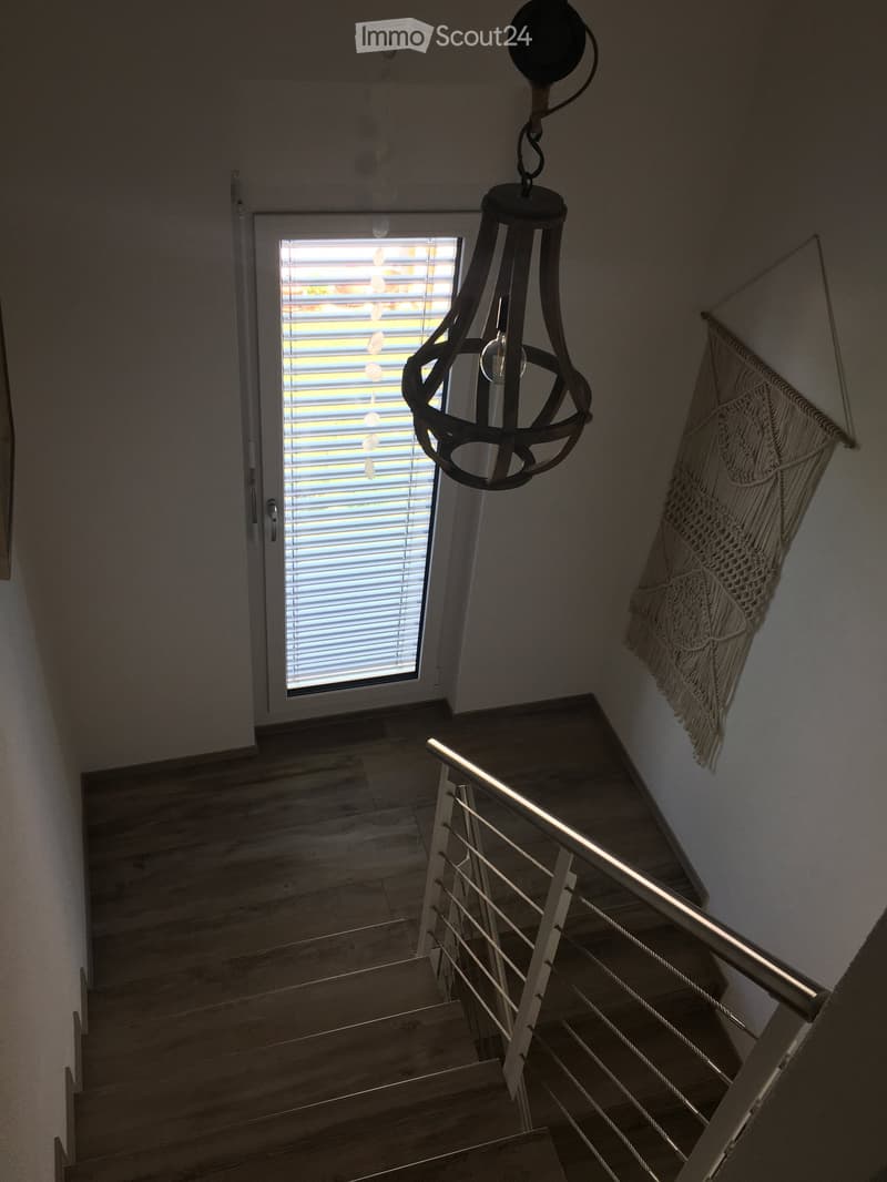 Staircase with wooden steps, metal railing, and a hanging pendant light. A window with blinds is visible at the top of the stairs, and a woven wall hanging is on the right wall.