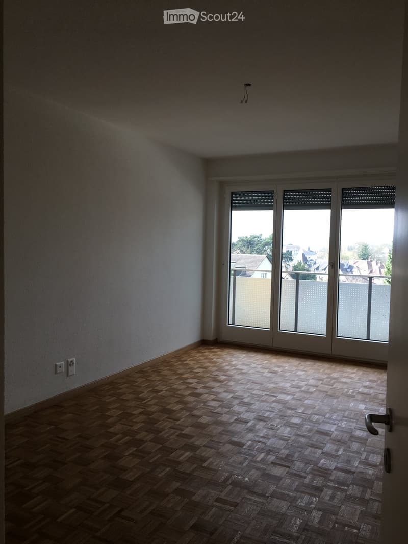 Empty room with wooden herringbone flooring, white walls, and large sliding glass doors leading to a balcony with a view of the neighborhood.
