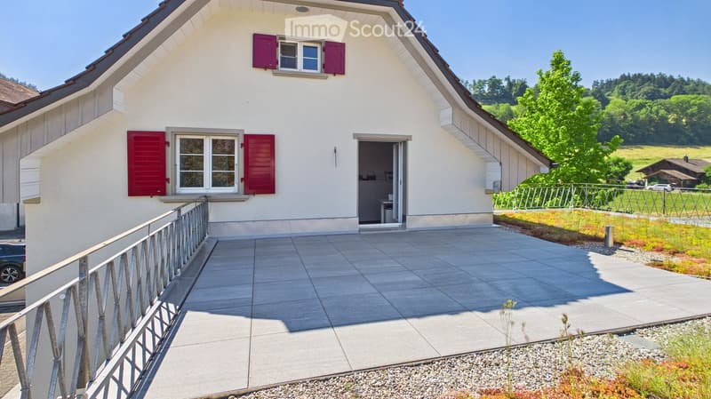 Two-story house with light beige walls, red shutters, and a tiled patio area. The house features a balcony with a metal railing and a small garden area with gravel and plants.
