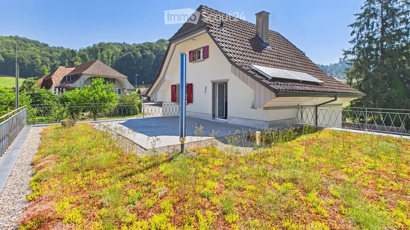 A modern two-story house with a tiled roof, white exterior, and red shutters. The house features a raised ground floor with a green roof and a balcony area.