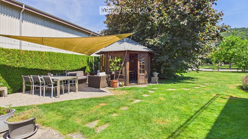 Well-maintained garden with a seating area, including a table, chairs, sofa, ottoman, and a small wooden gazebo with a bar area. The space is shaded by a large canopy and surrounded by greenery.
