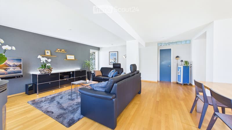 Modern living room with light wooden flooring, grey walls, and a large window. Features a black leather sofa, coffee table, TV stand, dining table with chairs, and decorative items including a flower vase and picture frames.