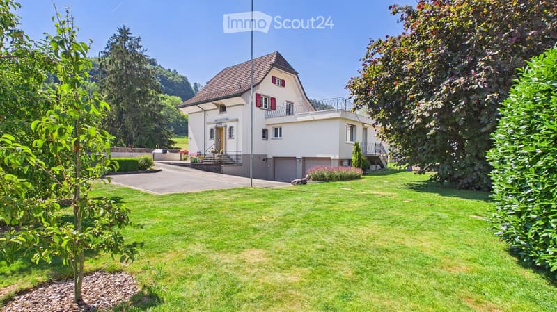 Two-story house with a red-tiled roof, white walls, and red shutters. Features a garage, a well-maintained lawn, and a small balcony on the upper floor.