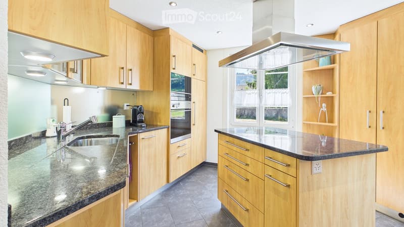 Modern kitchen with light wooden cabinets, granite countertops, stainless steel appliances, and a central island with a range hood.