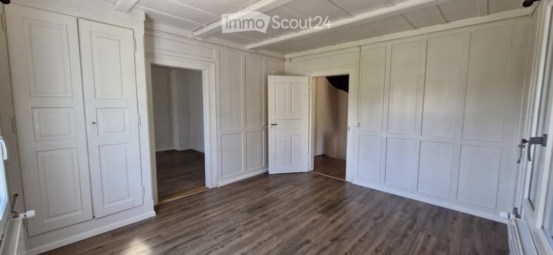 Empty room with white paneled walls, wooden flooring, and multiple doors leading to other spaces.