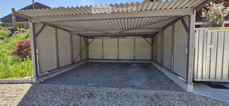 Carport with multiple garage doors, covered by a metal roof, surrounded by greenery and gravel.