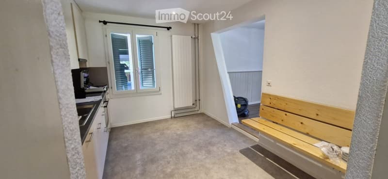 A small, unfurnished room with a wooden bench, a kitchen area with cabinets and a stove, and a window with shutters.