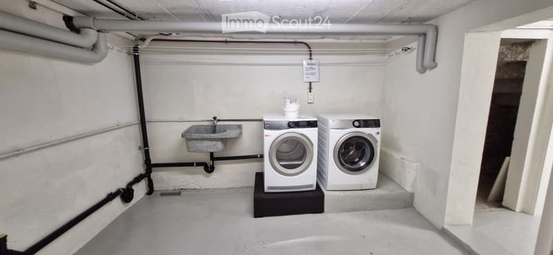 Utility room with a washing machine, dryer, and sink, featuring exposed pipes and a concrete floor.