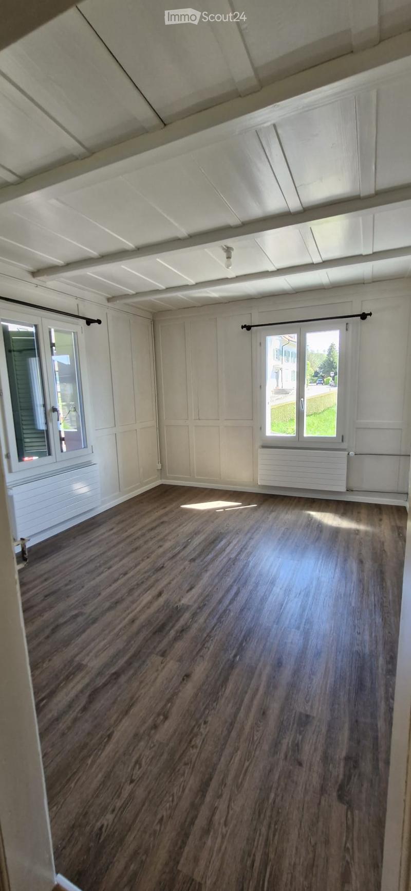 Empty room with wooden flooring, white walls, large windows, and a sliding door leading outside.