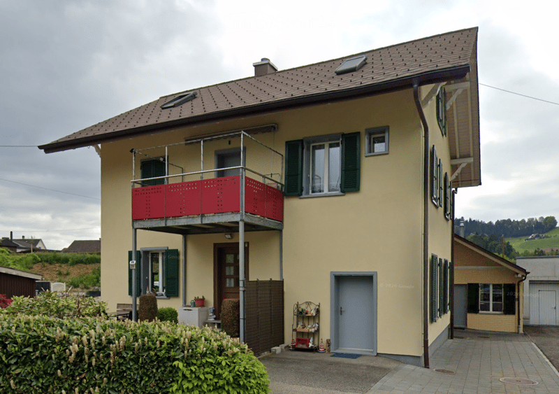 Two-story house with a balcony, green shutters, and a small garden area.