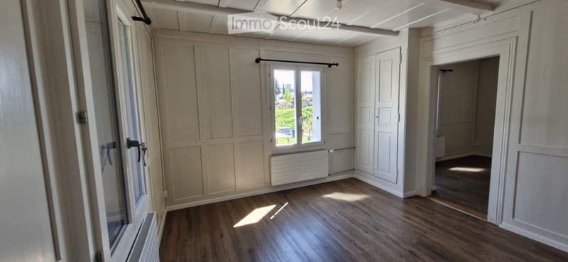 Empty room with wooden flooring, white walls, and large windows allowing natural light. The space features built-in closet doors and a radiator under one window.