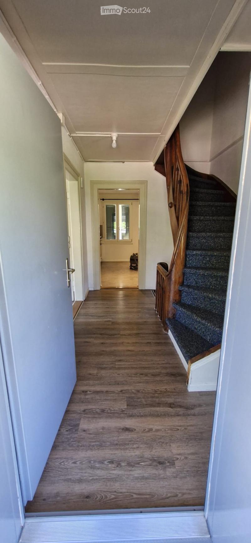 A long hallway with wooden flooring, white walls, and a staircase with a wooden railing leading upwards. A door on the left opens to another room, and a small window is visible at the end of the hallway.
