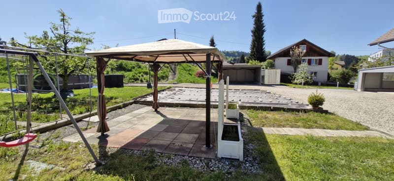 A well-maintained garden with a patio, swing, small fountain, and surrounding greenery.