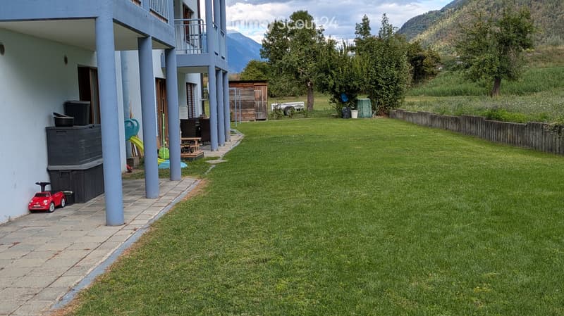 Lush green lawn with a paved pathway leading to a house with blue columns and a balcony. A small wooden shed and a few outdoor items are visible near the house.