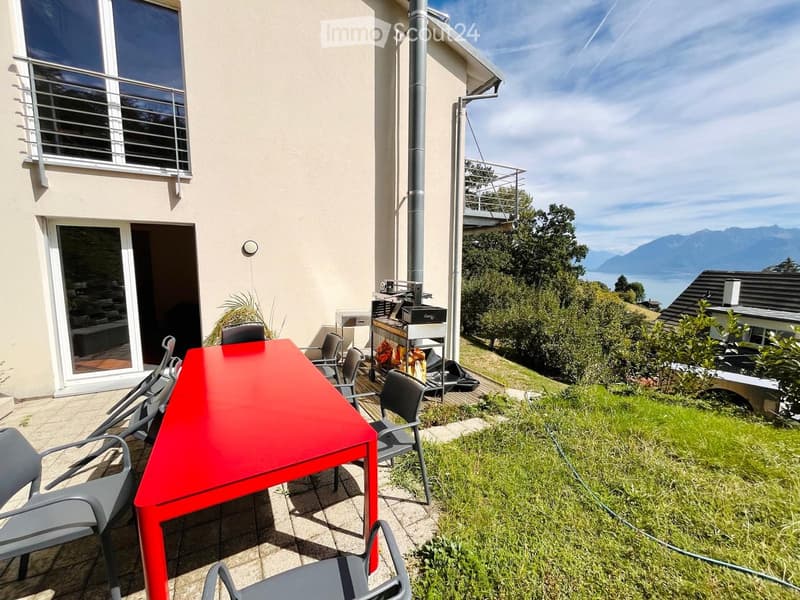 Outdoor patio with a red table and several chairs, overlooking a scenic view of mountains and a lake.