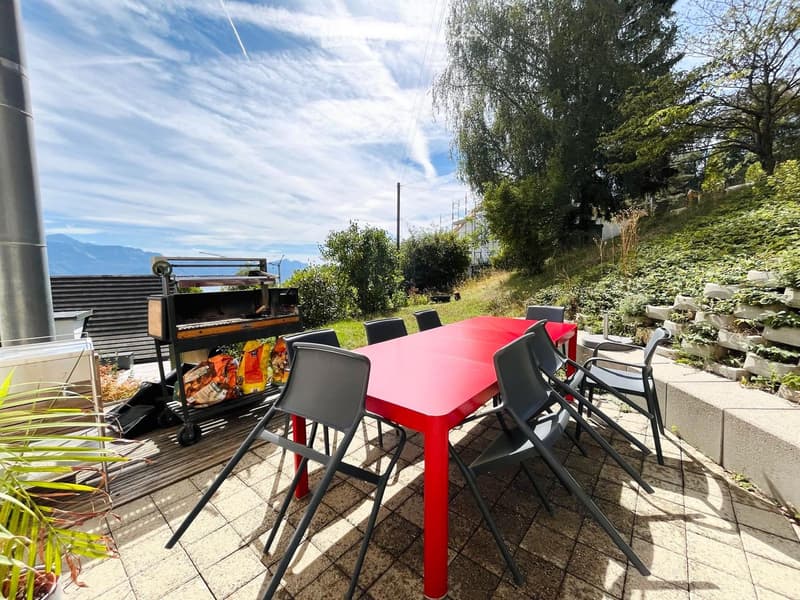 Outdoor patio with a red table, black chairs, and a grill, surrounded by greenery and a scenic mountain view.