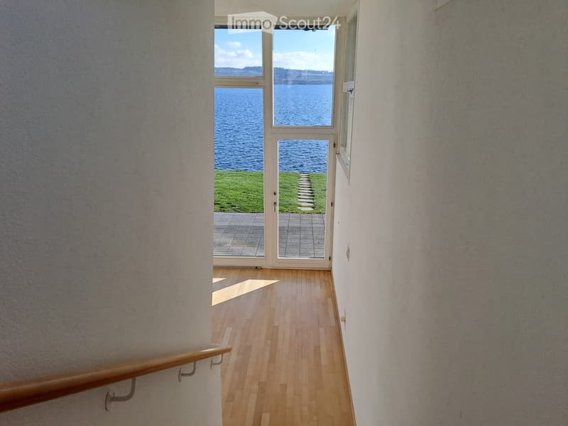 Empty corridor with a glass door leading to a lake view, concrete floor, wooden handrail