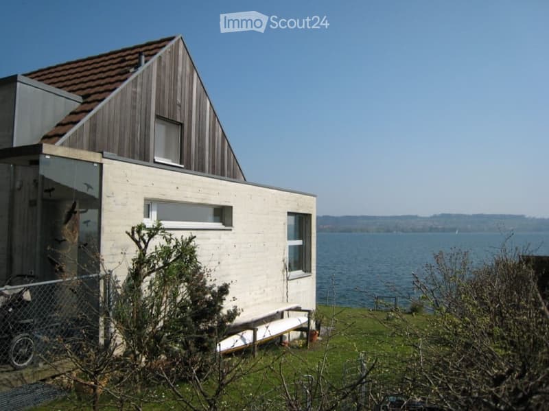 A modern house with a red tiled roof, a glass wall, a white bench in front, a large water body nearby, and a green field.