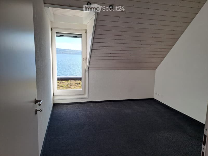 Empty room with grey carpeted floor, white walls, and a large window offering a view of the water. The ceiling has wooden paneling and recessed lighting.