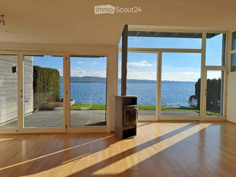 Open living space, large windows with lake view, fireplace, wooden floor, sliding glass doors.