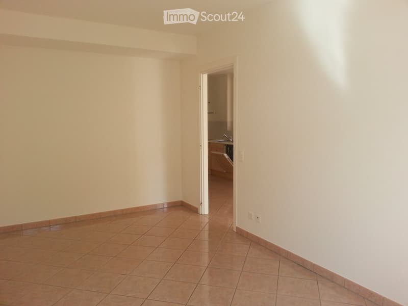 Empty room with tiled floor and white walls, featuring an open doorway leading to a small kitchen area with cabinets and a sink.