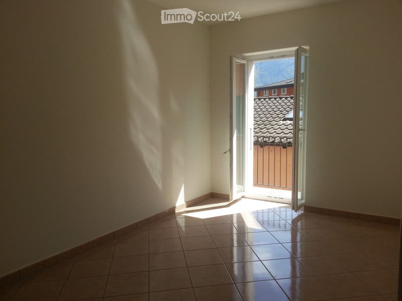 Empty room with tiled floor and open sliding glass door leading to a balcony area with a view of a tiled roof and a building.