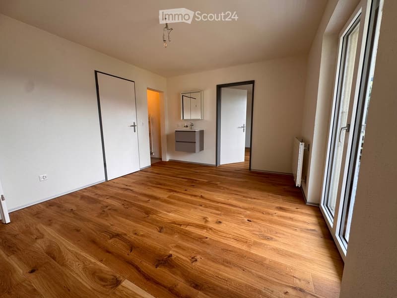 Empty room with wooden flooring, white walls, and large windows. Features a small bathroom area with a sink and cabinet visible in the background.