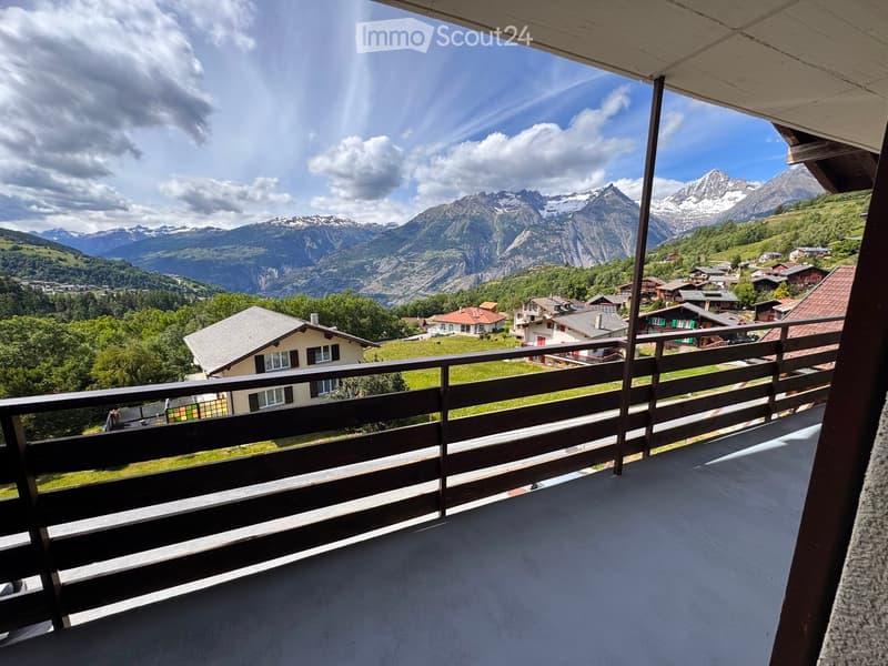 Balcony with wooden railing overlooking a scenic mountain view and a small village with houses and greenery.