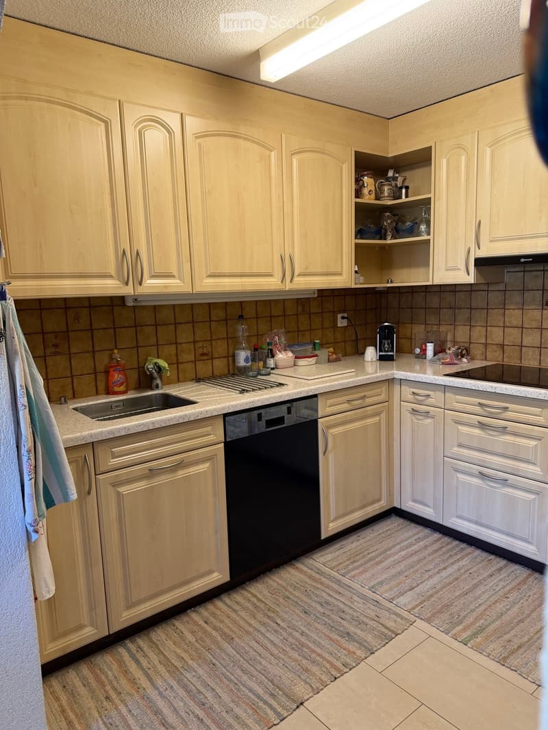 A well-lit kitchen with light wooden cabinets, a stainless steel sink, a dishwasher, and a countertop with various items including a coffee maker, bottles, and cups. The floor is tiled, and there is a small rug in the center.