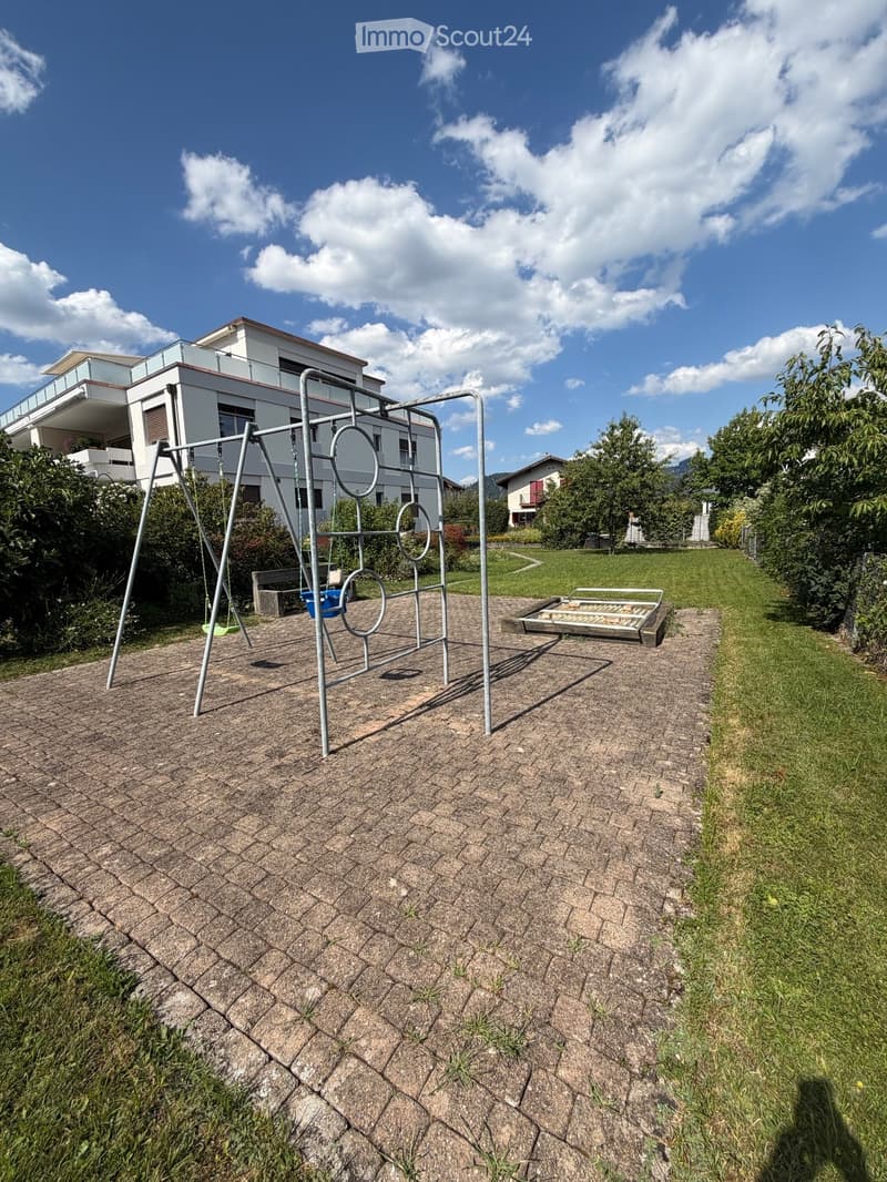 Outdoor playground area with a swing set, monkey bars, and a small bench, surrounded by a paved path and grassy lawn, with residential buildings in the background.