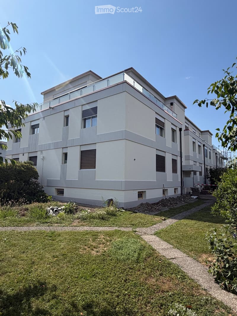 Modern two-story building with white facade, large windows, and a rooftop terrace. The surrounding area includes a well-maintained lawn, gravel pathways, and some greenery.