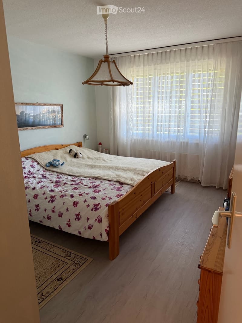 Cozy bedroom with a wooden bed, floral bedspread, white curtains, a hanging lamp, and a small rug on the floor.