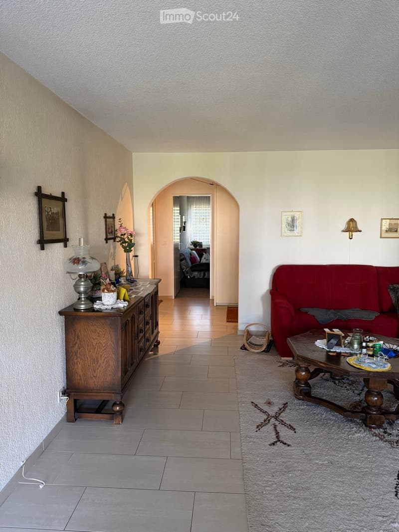 Living room with a red sofa, wooden coffee table, sideboard, table lamp, vase with flowers, and picture frames on the walls.