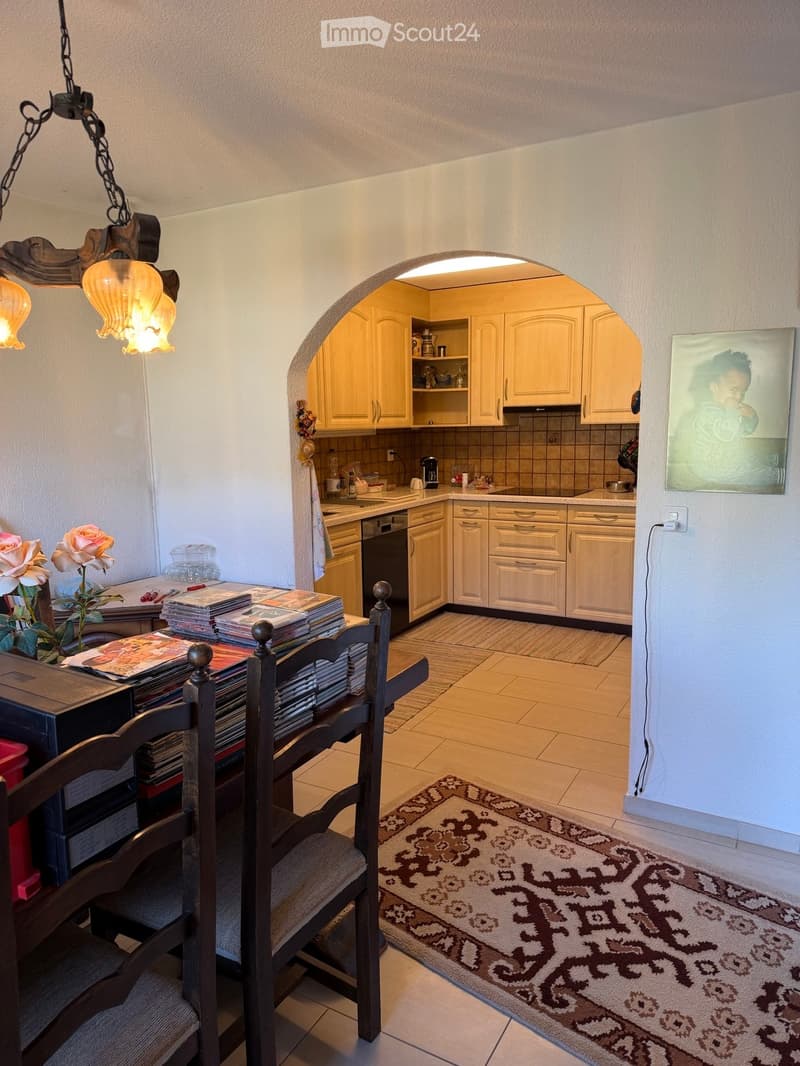 A cozy dining area with a wooden table and chairs, a flower vase, and a stack of CDs. An adjacent kitchen features light-colored cabinets, a dishwasher, and a sink.