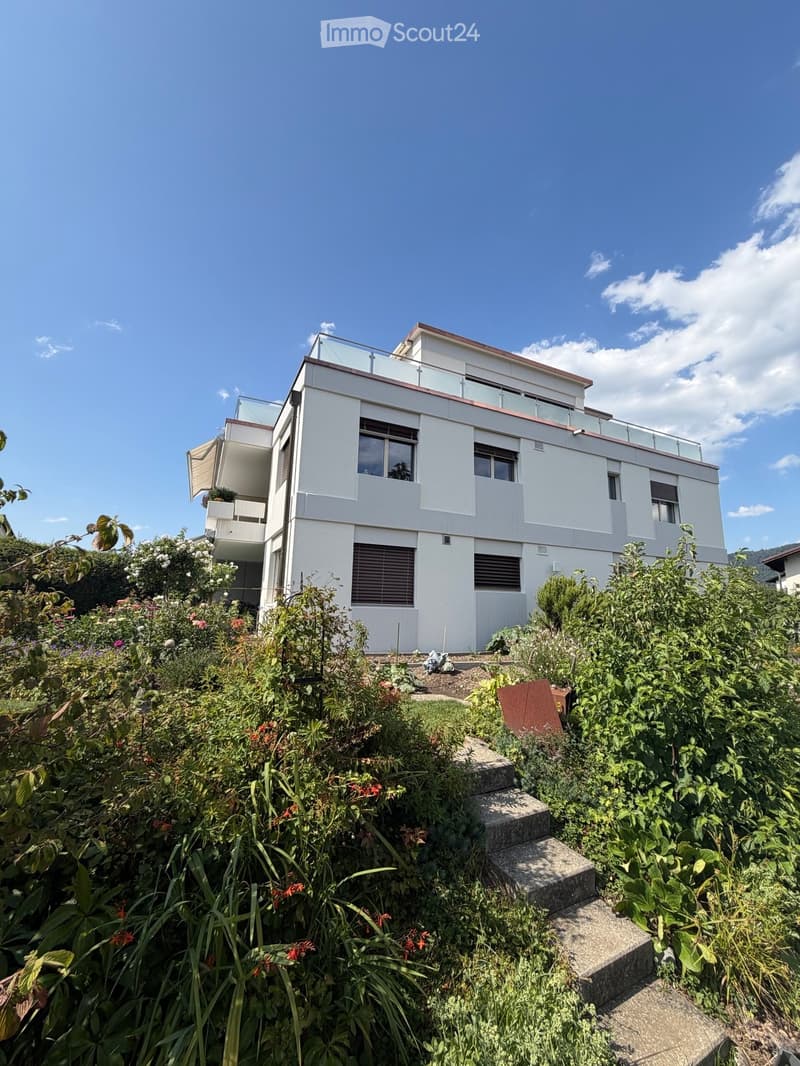 Modern two-story house with white exterior, large windows, and a rooftop terrace surrounded by lush greenery and a staircase leading up.
